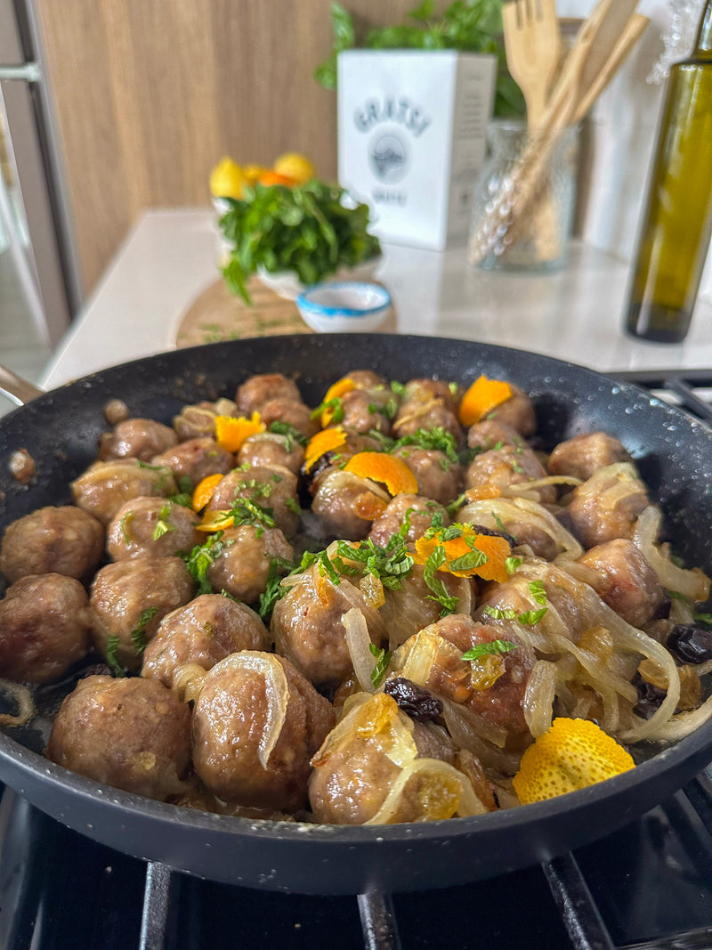 Meatballs cooking in a black skillet with sliced onions, orange peel, and herbs, set on a stovetop in a modern kitchen with fresh ingredients in the background.