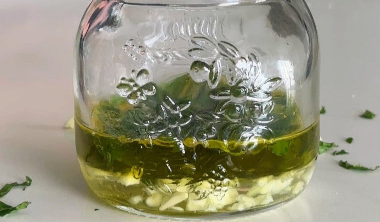 A glass jar with chopped garlic, olive oil, and herbs sits on a white surface. Some herb pieces are scattered around the jar. The jar has a decorative floral pattern embossed on it.