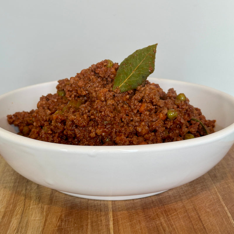 A white bowl filled with spiced ground meat garnished with a bay leaf, placed on a wooden surface against a plain light background.