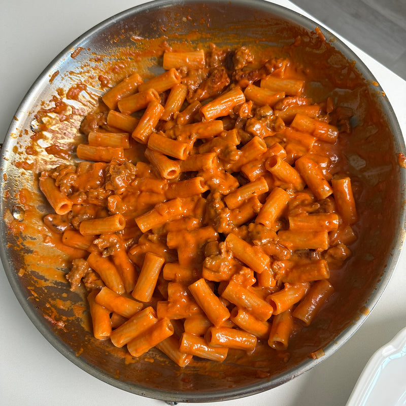 A skillet filled with rigatoni pasta coated in a creamy red tomato sauce, with bits of ground meat mixed in. The dish sits on a white surface with some sauce smeared on the sides of the pan.