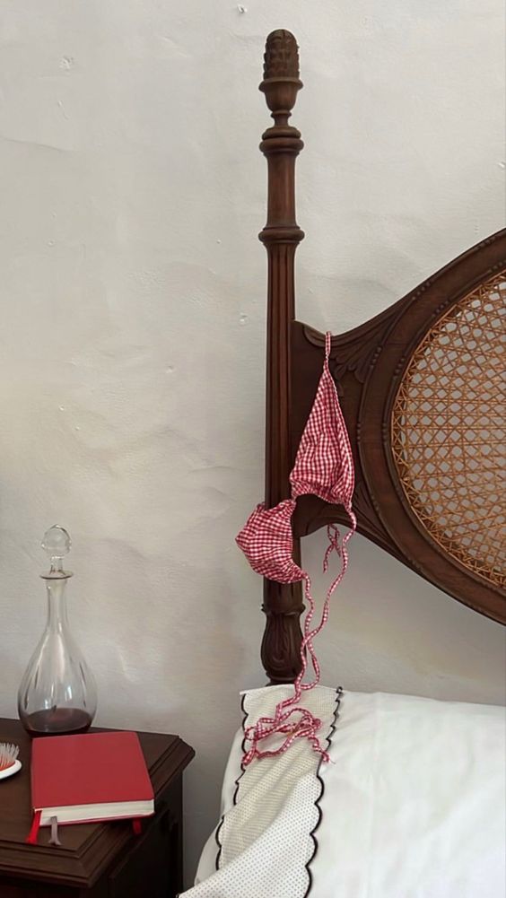 A red gingham bikini top hangs on the wooden post of a vintage bed. Next to the bed, a nightstand holds a glass decanter, a closed red book, and a small dish. A white pillow with black trim is on the bed.