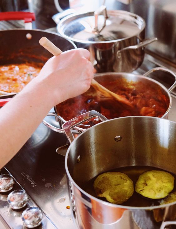 A person stirs a pot of red sauce on a stove with a wooden spoon, while another pot in the foreground contains two pieces of zucchini boiling in water.