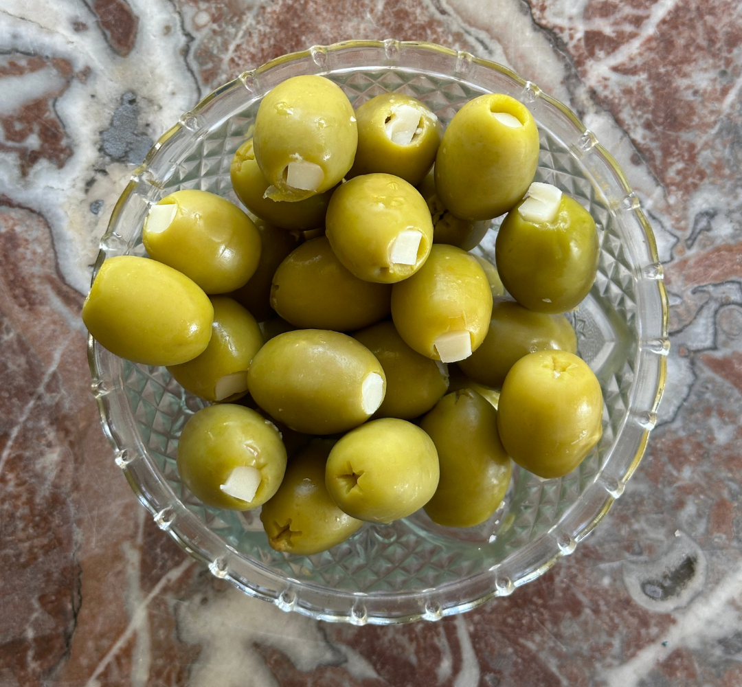 A glass bowl filled with green olives, some of which are stuffed with small pieces of garlic, sits on a marbled surface.