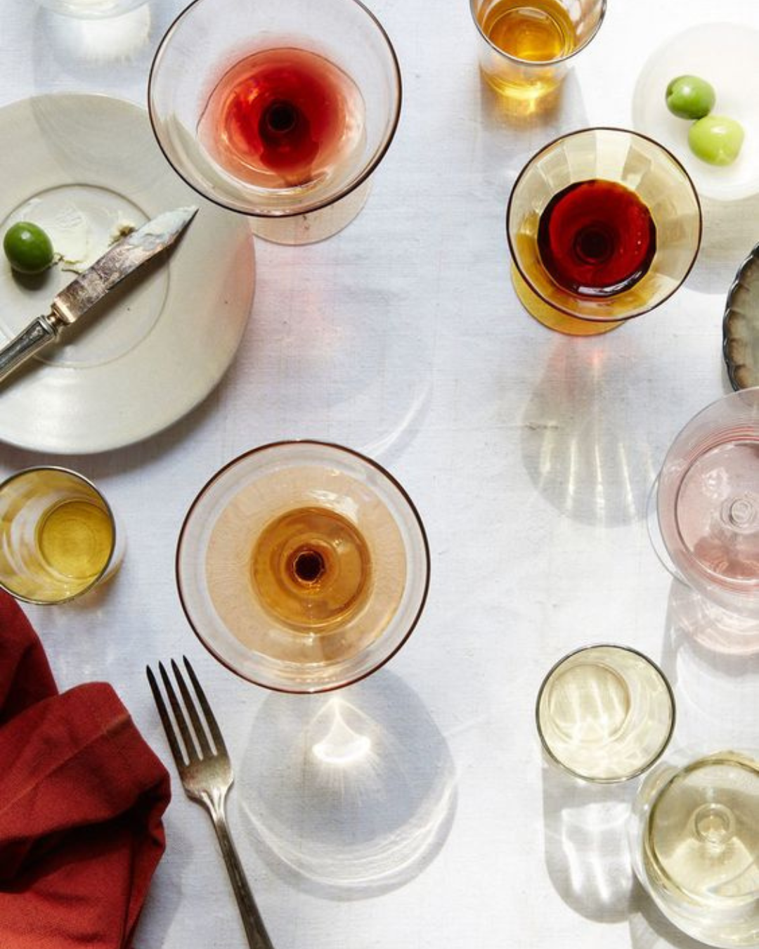 A table set with empty and half-filled glasses of various colored drinks, an empty plate with a knife and green olive, a fork, a red napkin, and scattered green olives, all on a white tablecloth.
