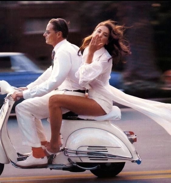 A man and woman ride a white scooter; the man drives while the woman, in a flowing white outfit, sits behind him blowing a kiss. Both appear joyful, with motion blurring the background.