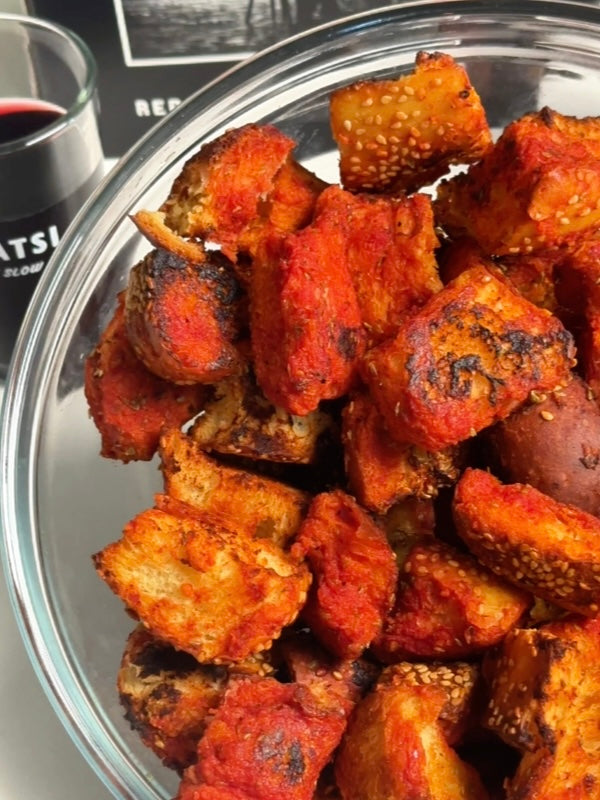 A glass bowl filled with crispy, golden-red bread cubes coated in spices and sesame seeds, with a glass of red wine and a blurred background.