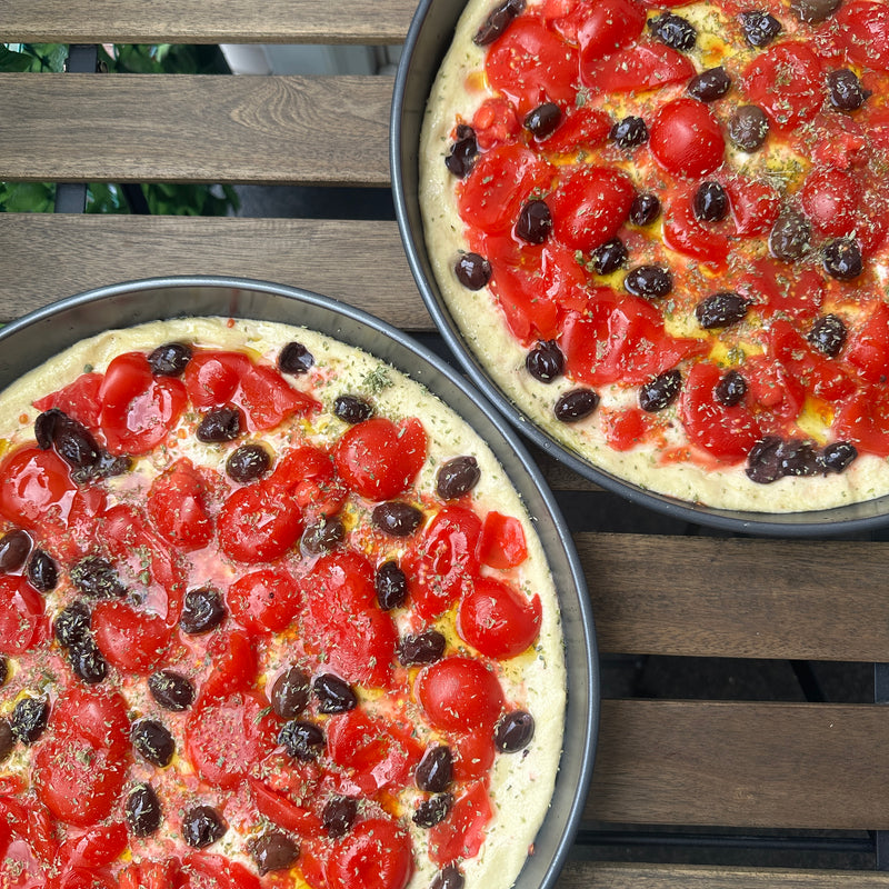 Two round baking pans filled with focaccia dough topped with chopped cherry tomatoes, black olives, and herbs, placed on a wooden table.