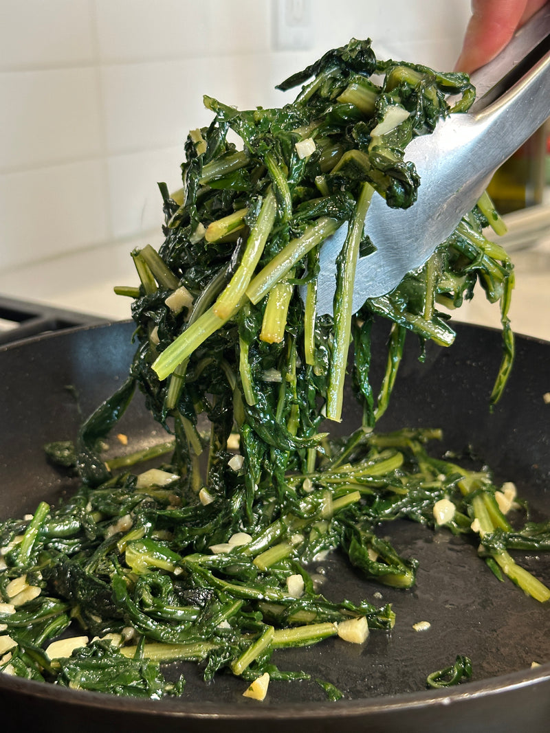 A close-up of sautéed spinach with garlic being lifted from a black pan using metal tongs. The greens look tender and glossy, with chopped garlic pieces visible. The background features a tiled white kitchen wall.