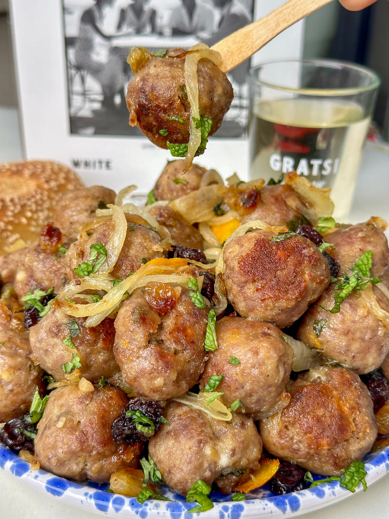 A close-up of a plate of browned meatballs garnished with cooked onions, herbs, and raisins. A hand holds a wooden fork with a meatball above the plate. A glass of white wine and a bun are in the background.