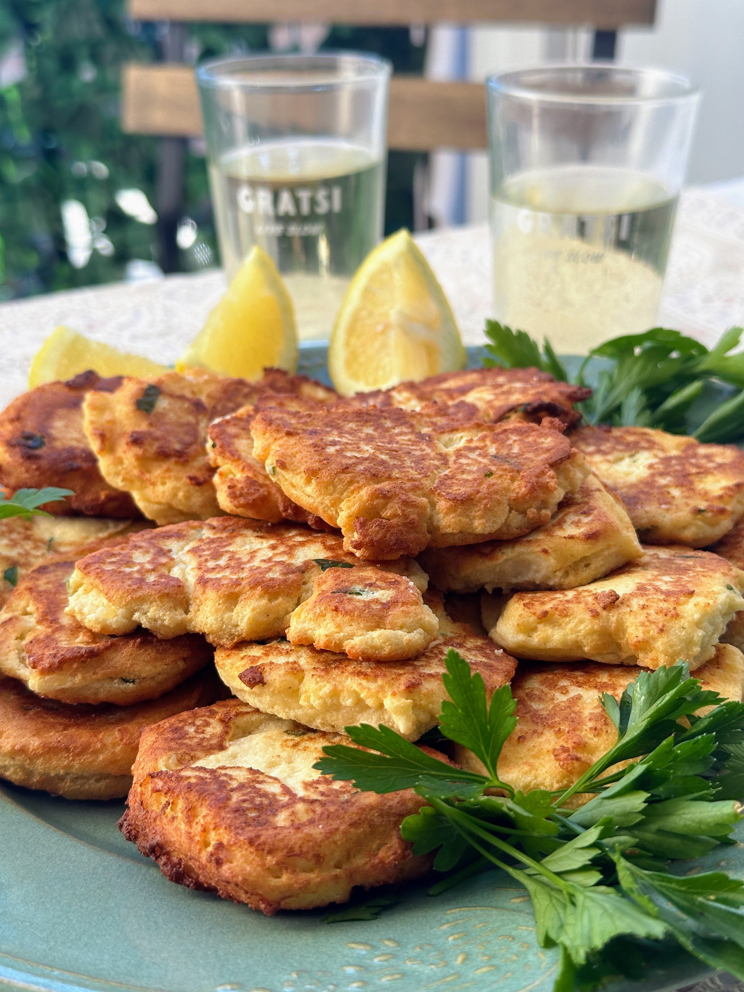A plate of golden, crispy fritters garnished with fresh parsley and lemon wedges, with two glasses of white wine in the background on an outdoor table.