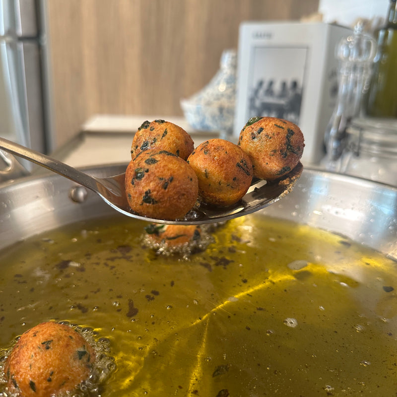 A close-up of a metal spoon holding several golden-brown, round fritters with herbs, above a pan of hot oil on a stovetop. More fritters are seen frying in the oil below.