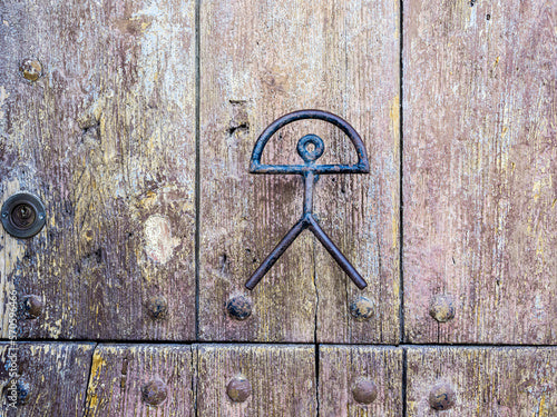 A weathered wooden door with metal studs features an abstract black metal symbol resembling a stick figure with a semicircle above its head.