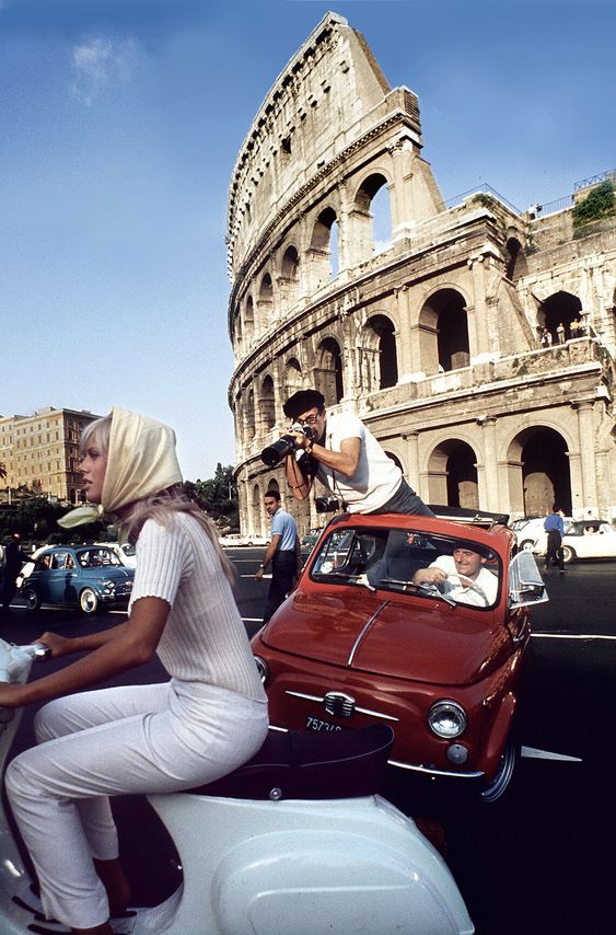 A woman in a white outfit and headscarf rides a scooter past the Colosseum in Rome. A man stands through the sunroof of a red car, taking photos, while another person drives. Several vintage cars are in the background.