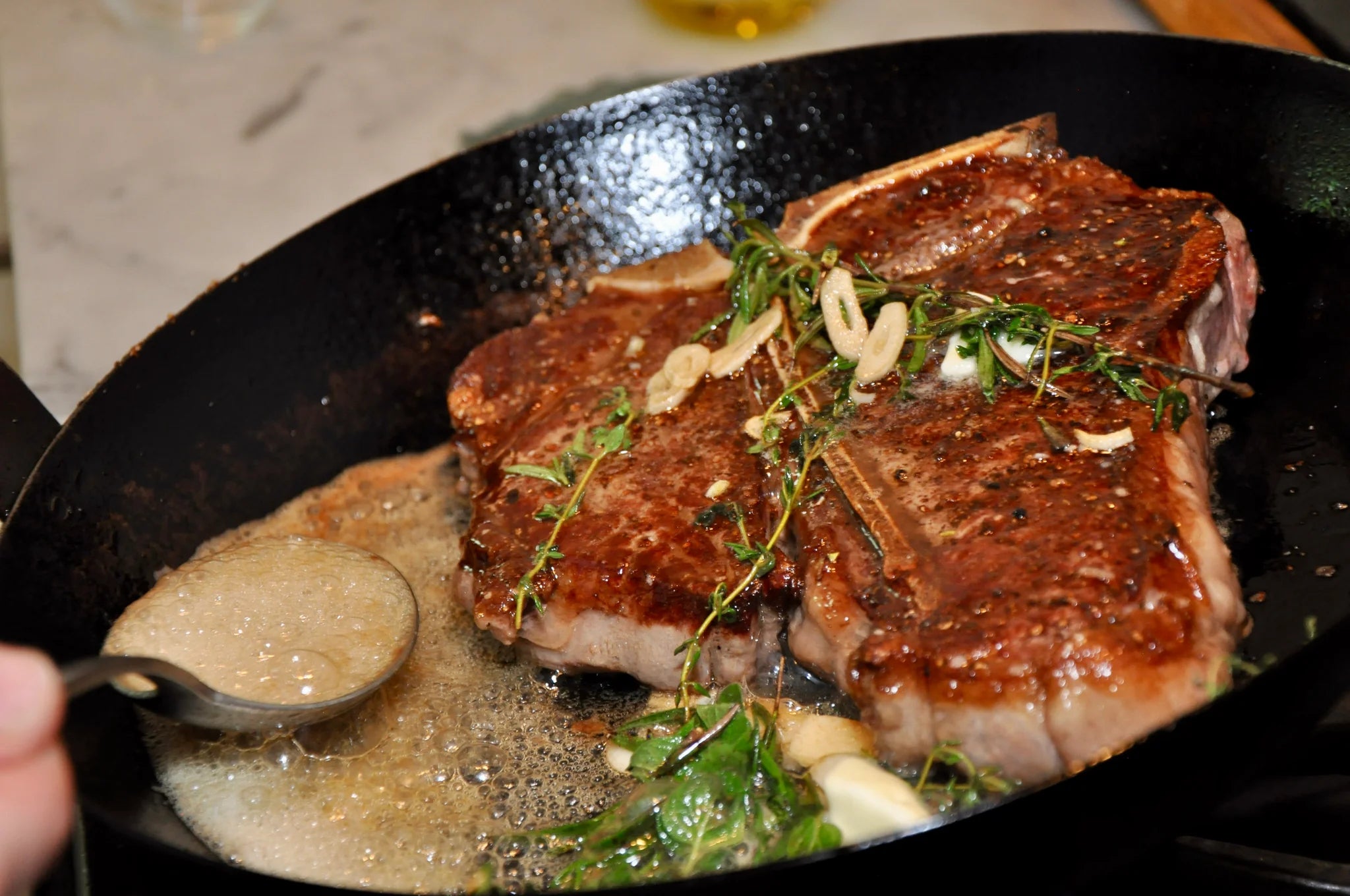A steak cooking in a cast iron pan with herbs, garlic, and bubbling butter, as someone spoons the butter over the meat.