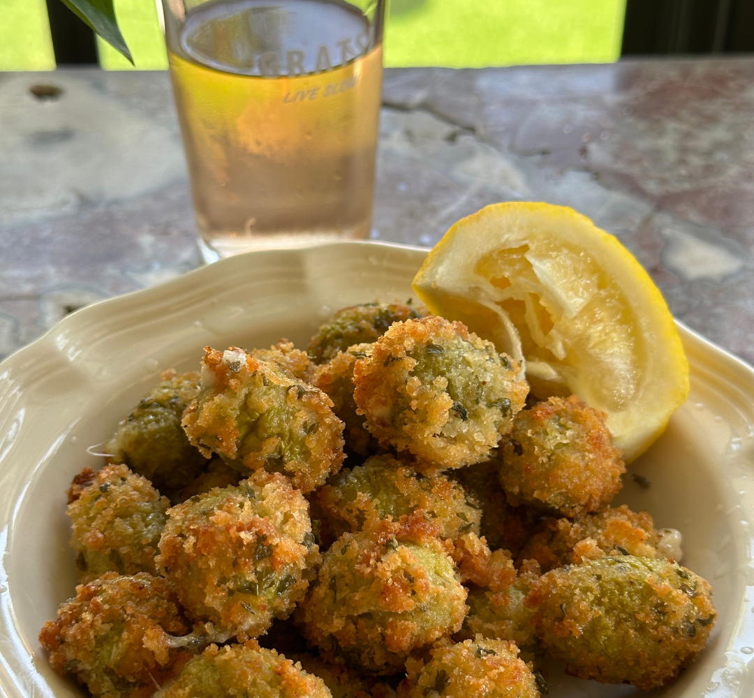 A plate of crispy, breaded fried olives garnished with a lemon wedge sits on a marble table. In the background, there is a glass of light-colored drink.