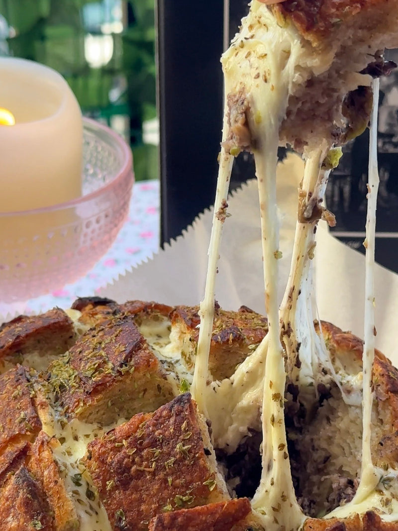 A close-up of pull-apart bread with a golden crust, topped with herbs and filled with gooey, stretchy melted cheese being lifted, with a lit candle in the blurred background.