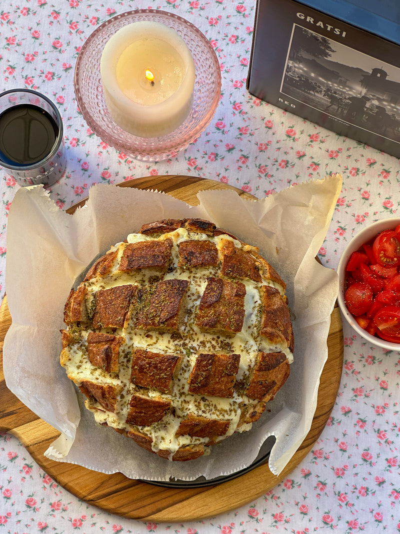 A round loaf of bread scored in a crisscross pattern, filled with melted cheese and herbs, sits on parchment paper atop a wooden board. Nearby are a candle, a glass of red wine, and a bowl of sliced cherry tomatoes.