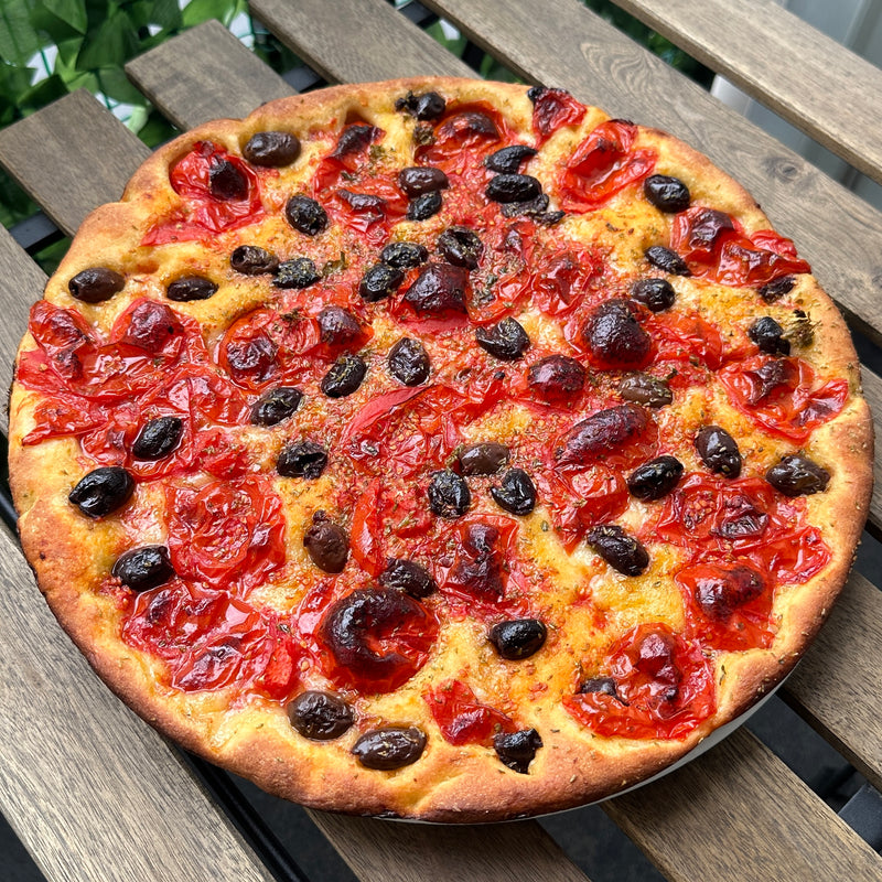 A freshly baked focaccia topped with halved cherry tomatoes, black olives, and herbs, sits on a wooden table outdoors. The crust is golden and slightly crisp, with a rustic and appetizing appearance.