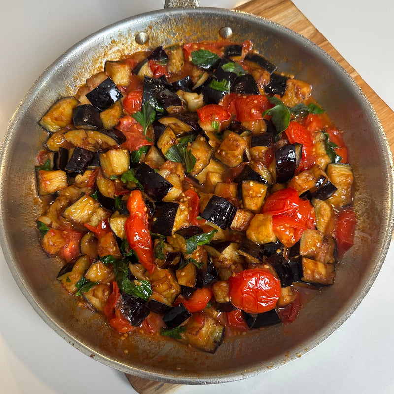 A skillet filled with sautéed eggplant, cherry tomatoes, and fresh basil in a rich tomato sauce, resting on a wooden board.