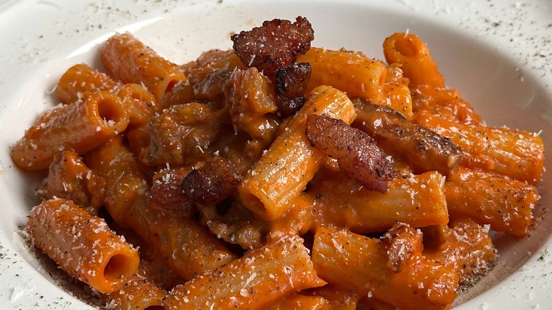 A close-up of rigatoni pasta coated in a creamy tomato sauce, topped with crispy pieces of bacon and grated cheese, served on a white plate with a sprinkle of black pepper.