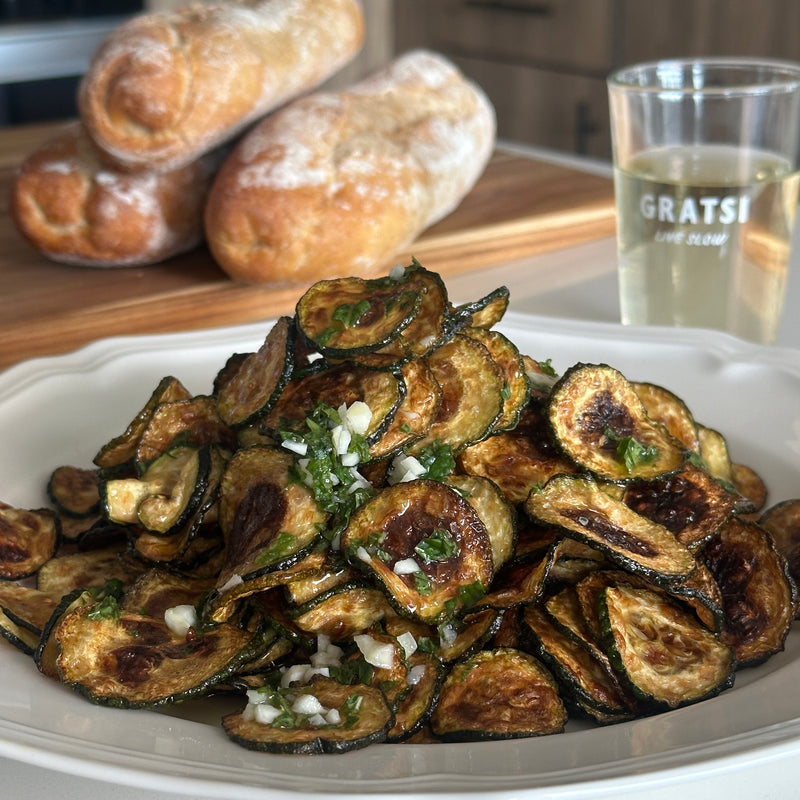 A plate of crispy fried zucchini slices garnished with diced garlic and herbs sits on a white dish, with two loaves of bread and a glass of white wine in the background.