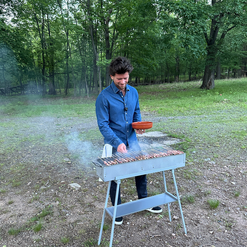 A man in a blue shirt grills skewers on a barbecue in a grassy outdoor area surrounded by trees. He is holding a bowl and smiling while tending to the food. Smoke rises from the grill.