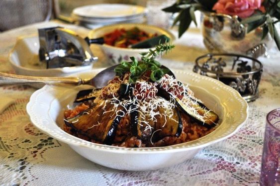 A bowl of pasta topped with grilled eggplant slices, tomato sauce, and grated cheese sits on a lace-covered table, surrounded by serving dishes and a floral centerpiece.