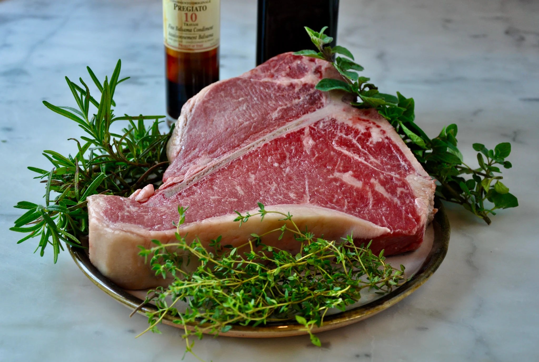 A raw T-bone steak sits on a plate surrounded by fresh herbs, including rosemary and thyme, with bottles of seasonings or oil in the background on a marble surface.