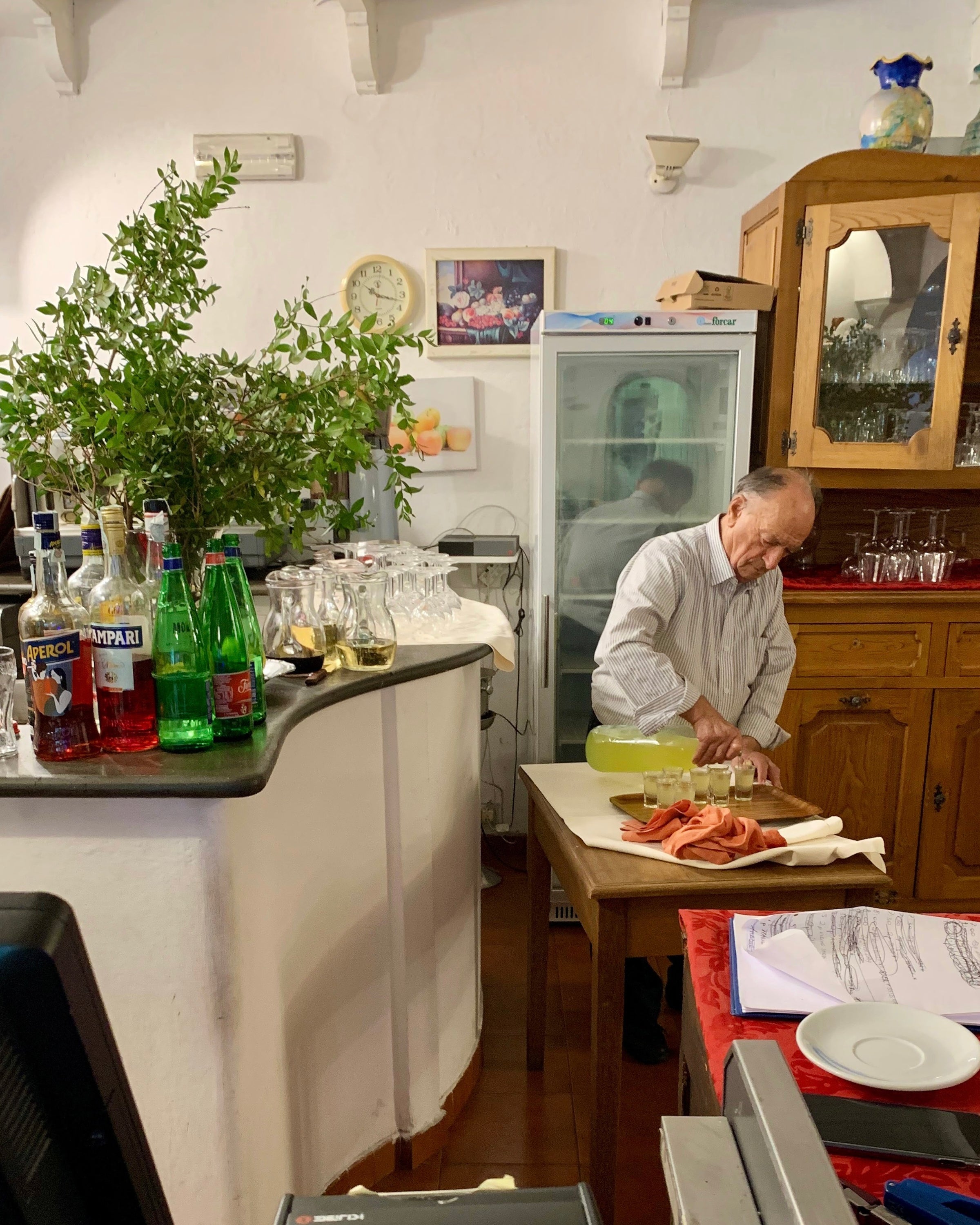 An elderly man in a striped shirt prepares food behind a counter in a cozy, rustic kitchen with bottles, glasses, a clock, and greenery on display. A refrigerator and wooden cabinet are in the background.