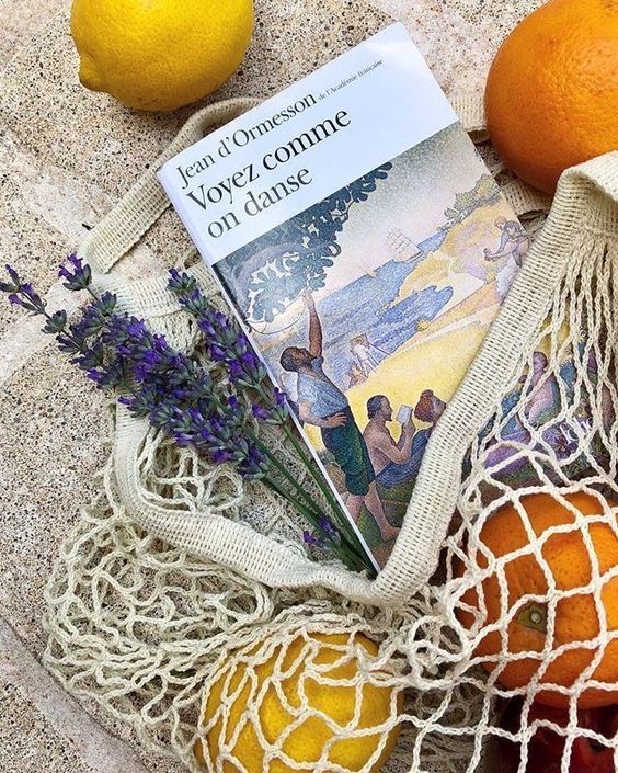 A mesh bag with oranges, a lemon, lavender sprigs, and the book Voyez comme on danse by Jean dOrmesson, placed on a light stone surface.