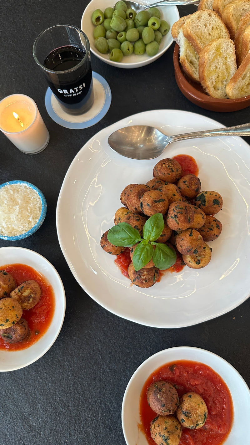 A plate of vegetarian meatballs in tomato sauce garnished with basil is on a table with bowls of grated cheese, green olives, toasted bread, a glass of red wine, and a lit candle.