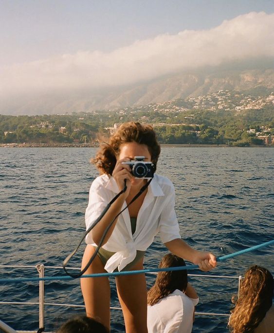 A woman on a boat, wearing a white shirt, takes a photo with a camera. The sea and coastline with mountains and trees are visible in the background under a partly cloudy sky. Other people sit nearby.