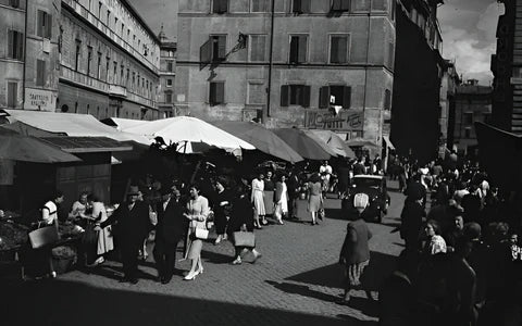 A busy outdoor market scene in a European city, with people shopping and walking among stalls under large canopies, surrounded by tall old buildings. The photo appears to be from the mid-20th century.