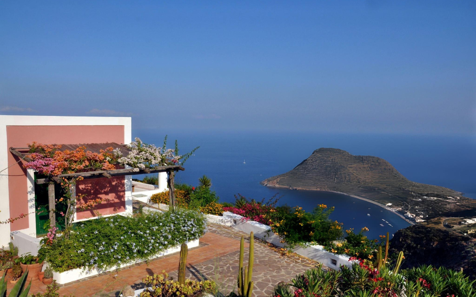 A terrace covered with flowering plants overlooks a deep blue sea and a small, hilly island in the distance, under a clear sky. Vibrant flowers and greenery surround the terrace, creating a peaceful coastal scene.