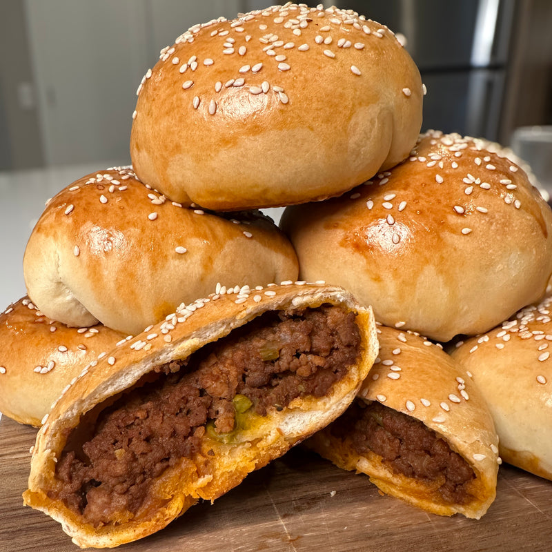 A stack of golden brown, sesame-topped baked buns, with one cut open to reveal a savory ground meat filling mixed with peas. The background shows a modern kitchen.