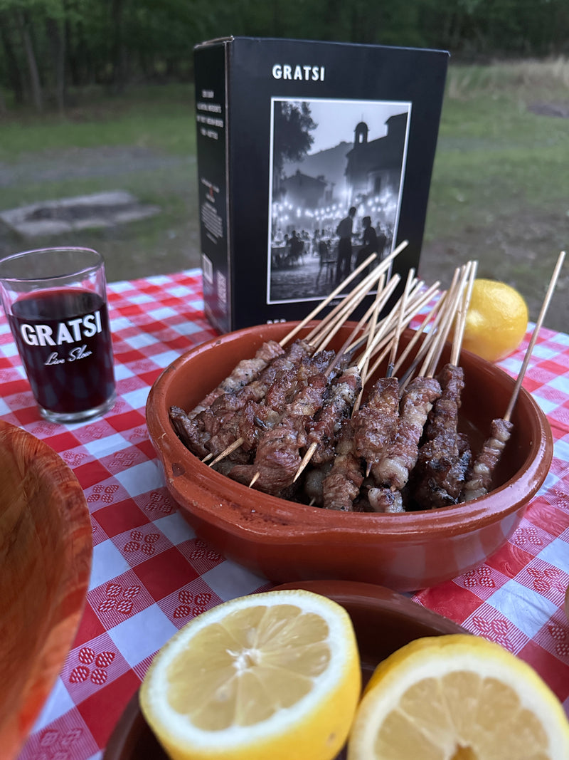 A bowl of grilled meat skewers sits on a red checkered tablecloth, next to halved lemons, a glass of red wine labeled GRATSI, and a black box with a vintage photo and GRATSI text on it. Outdoor setting in background.