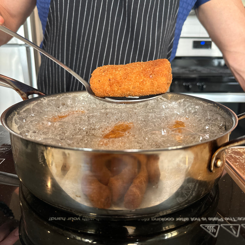 A person in a striped apron lifts a golden-brown breaded item from a pot of bubbling oil on a stovetop, with several more breaded items frying in the pot.