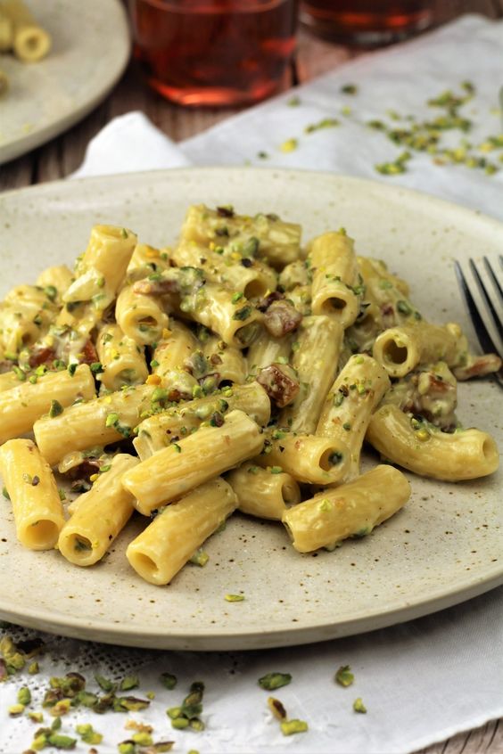 A plate of creamy rigatoni pasta garnished with chopped pistachios, served on a light-colored dish with a fork and a white napkin nearby.