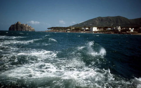 Waves crash in a blue sea near a coastline with white buildings and a green hill. A rocky island or cliff rises from the water in the distance under a clear, sunny sky.