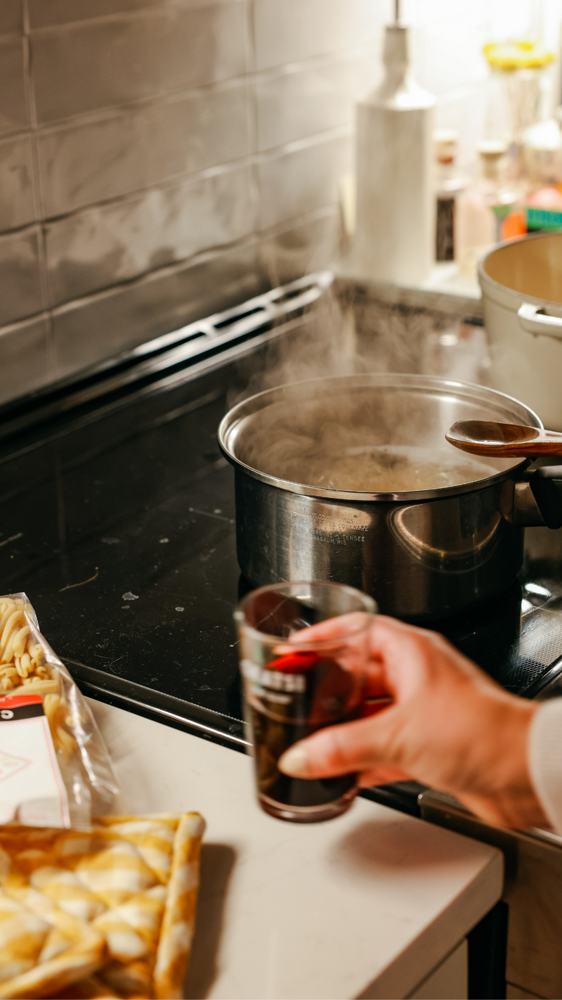 A person holding a glass of dark beverage stands by a steaming pot on a stovetop. A wooden spoon rests in the pot, and a package of pasta sits nearby on the counter. The kitchen is warmly lit.