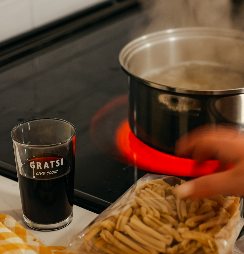 A glass of red wine labeled GRATSI LIVE SLOW sits on a counter near a stove with a pot of boiling water. A hand reaches for a bag of uncooked pasta in the foreground.