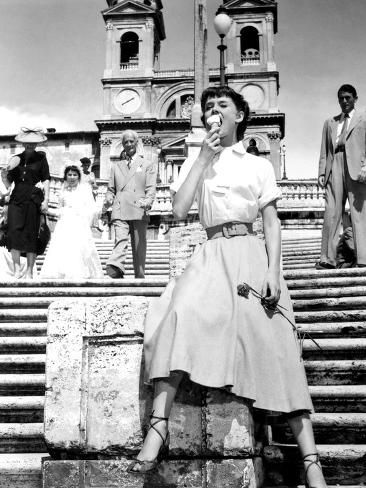 A woman in a mid-length skirt and blouse sits on stone steps, eating gelato, with a church and several people, including a bride and groom, in the background. The scene appears to be from the mid-20th century.