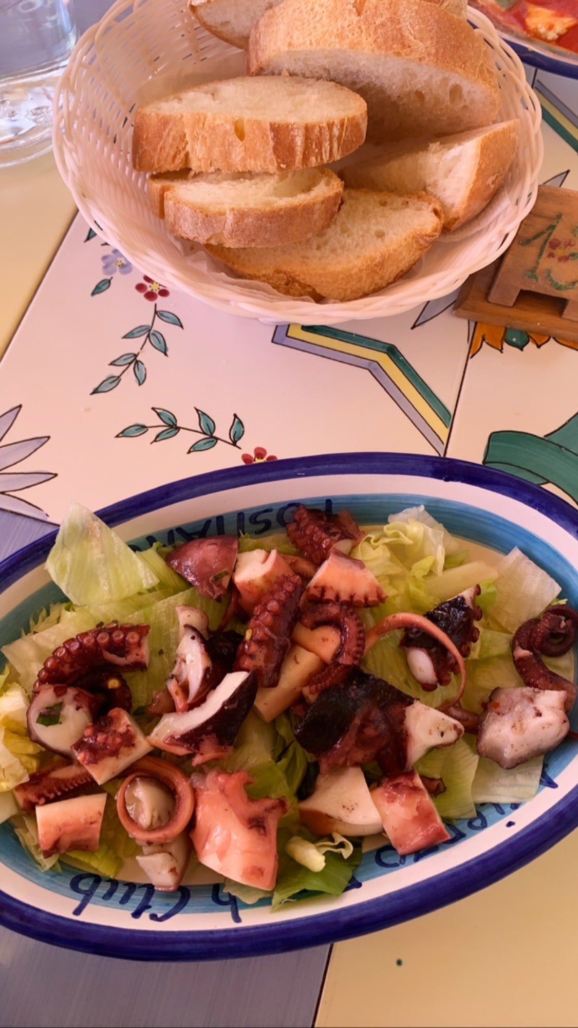 A plate of octopus salad with lettuce sits on a colorful table next to a basket of sliced bread.
