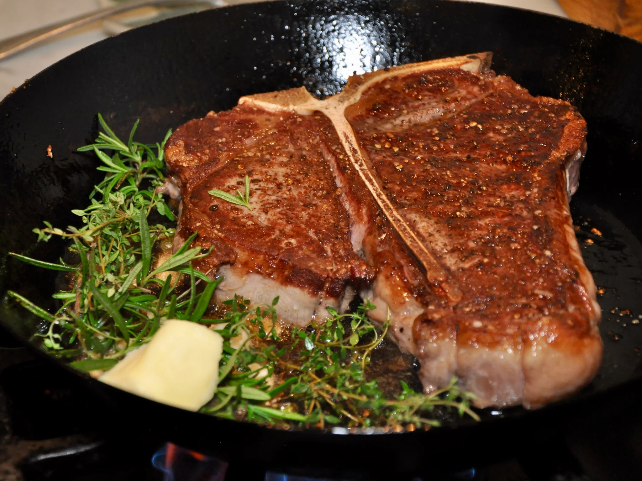 A seasoned T-bone steak is searing in a black cast iron skillet with sprigs of fresh rosemary, thyme, and a pat of melting butter.