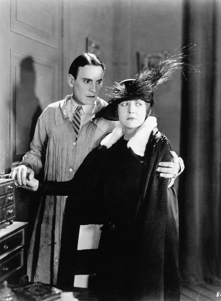 A man in an apron holds a womans arm as she stands in front of him, dressed in dark clothing with a large feathered hat. Both have serious expressions in this black-and-white vintage photograph.