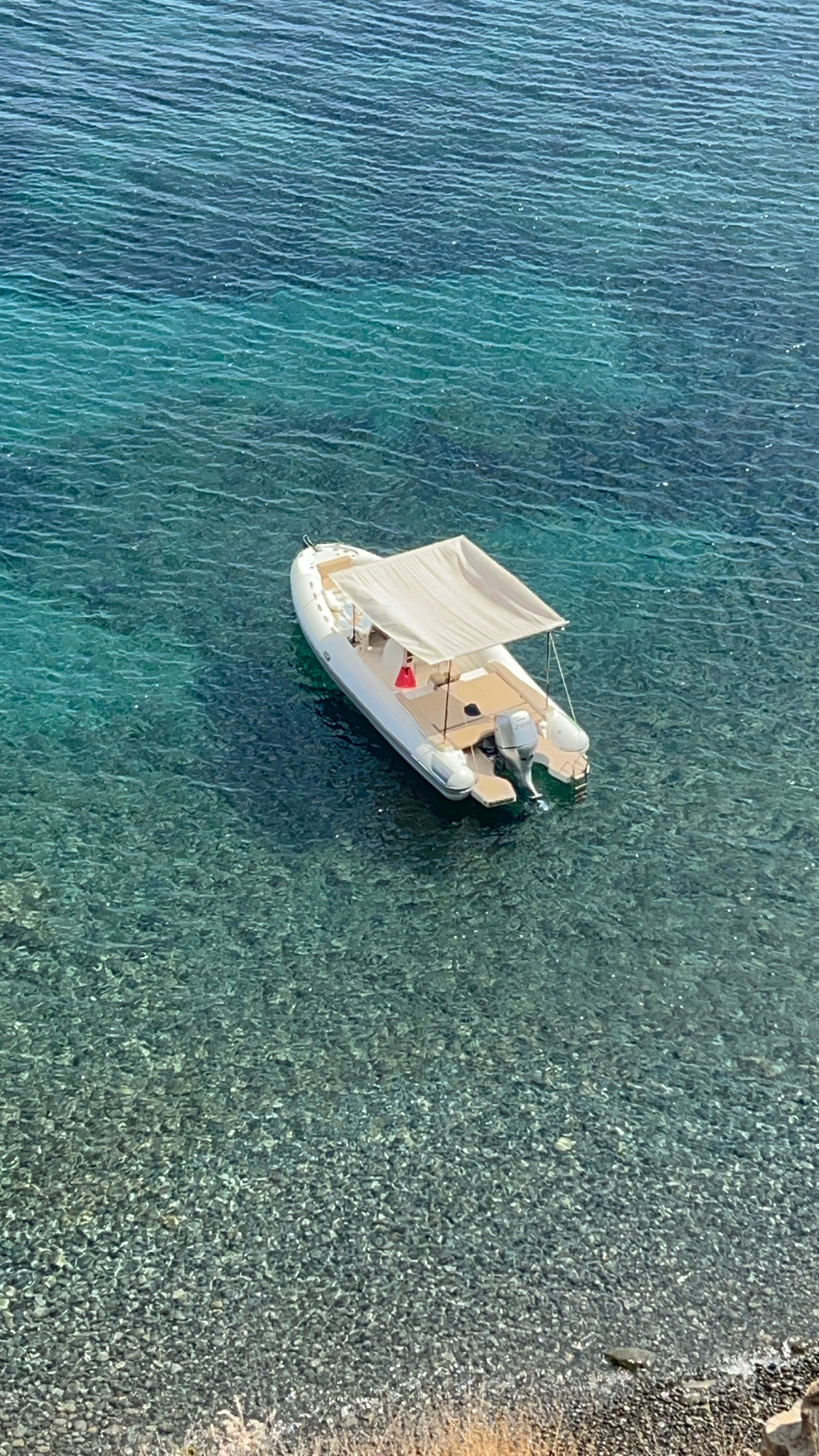 A small white boat with a beige canopy floats on clear turquoise water near a rocky shoreline, with the rocks visible through the shallow water below.