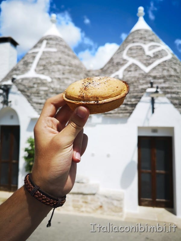 A hand holding a small pie or pastry in front of traditional white stone houses with cone-shaped roofs, decorated with white painted symbols, under a blue sky with clouds.
