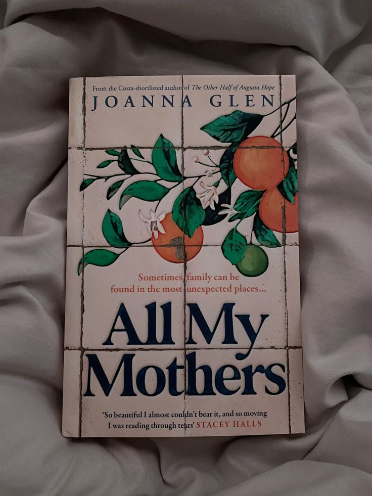 A book titled “All My Mothers” by Joanna Glen rests on rumpled white bedding. The cover features an illustration of oranges and white blossoms on green leaves with a quote and a review beneath the title.