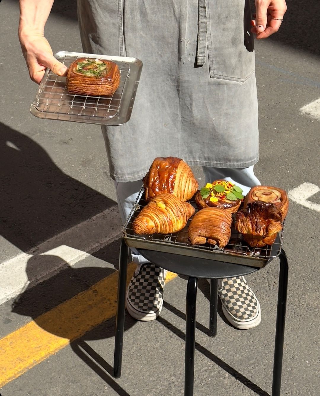 A person in a gray apron and checkered slip-on shoes stands on pavement, holding a tray with a pastry. Several assorted pastries rest on a rack placed over a black stool.