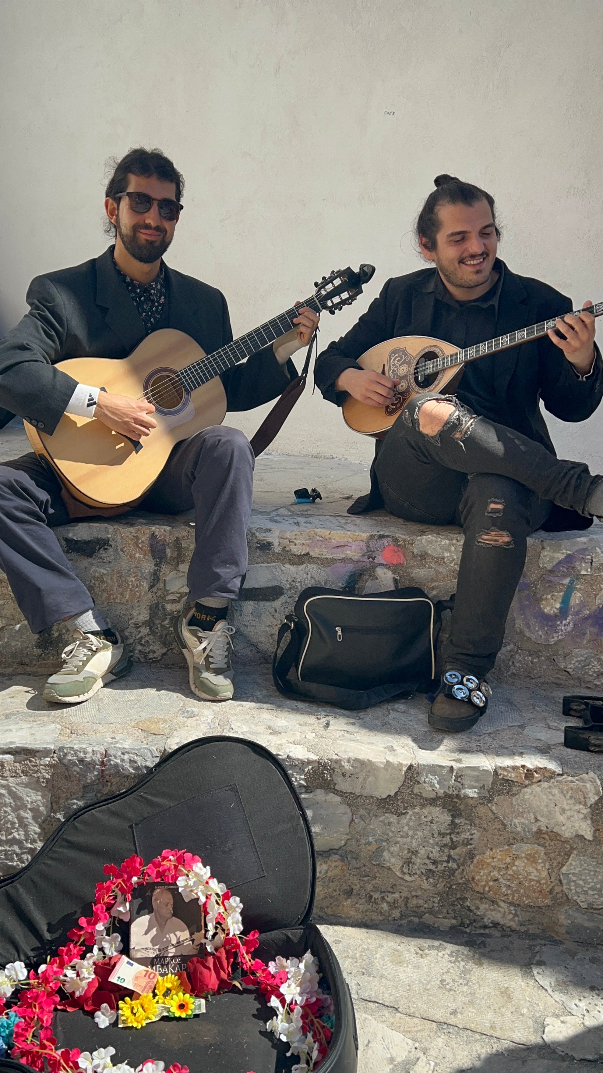Two young men sit on stone steps, playing stringed instruments and smiling. A guitar case with flowers, coins, and a photo is open in front of them, suggesting they are street performers. Sunlight casts shadows on the white wall behind them.